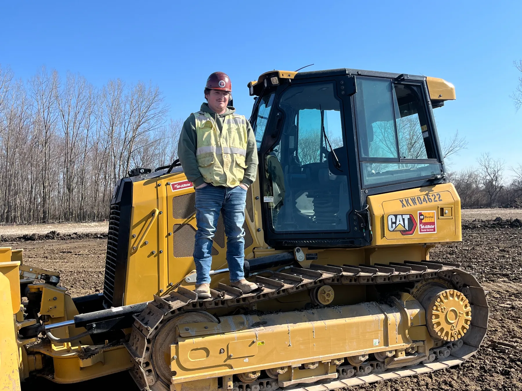 Matt Brooks in a hard hat and hi-vis safety vest standing on the track of a yellow CAT dozer on a dirt jobsite